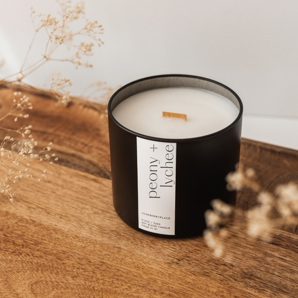 Black candle with a white label on a wooden surface with dried flowers.