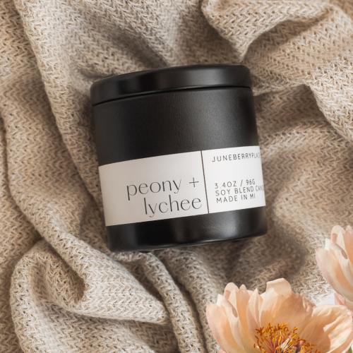 Black candle jar labeled 'peony + lychee' on textured fabric with flowers.